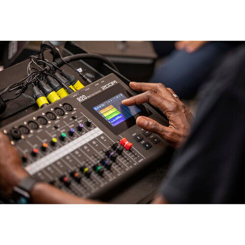 A person using the touch screen of the Zoom R20 audio mixer and recorder. The image shows the person's hand interacting with the colorful interface on the screen, which displays audio levels. Multiple input channels with connected cables are visible, emphasizing the device's multi-channel functionality. The setting appears to be a professional or home studio, highlighting the practical application of the equipment.