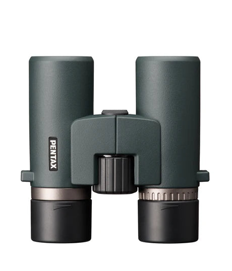 A top-down view of Ricoh Pentax AD 7x32 ED binoculars. The binoculars have a sleek, ergonomic design with a greenish matte finish. The central focusing wheel is positioned between the two barrels, with the Pentax branding visible on the left barrel. The eyepieces are capped, and the binoculars are designed for comfortable handling and ease of use, emphasizing durability and high-quality optics.