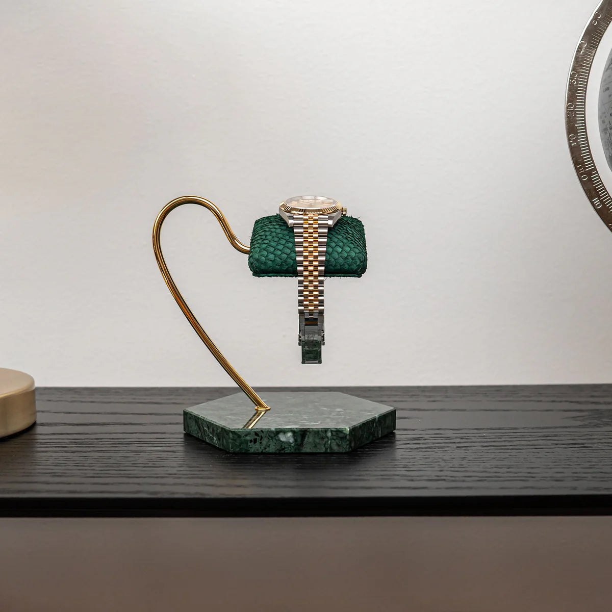 A luxurious gold wristwatch with a silver and gold metal band is displayed on a green textured cushion. The cushion is attached to an elegant gold stand with a green marble base, positioned on a dark wooden surface. The background is minimalist with a white wall, allowing the watch and stand to be the focal points of the image.