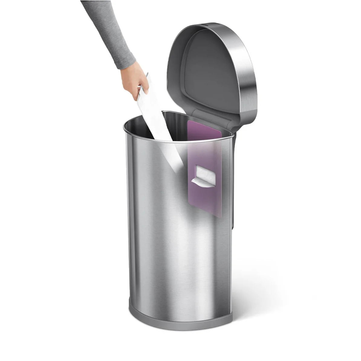 The image shows a Simplehuman touch-free sensor trash can with a person placing a white object (possibly a paper or a trash bag) into the can. The trash can has a stainless steel finish with a cylindrical shape. The lid is open, and the hand of the person is shown reaching into the trash can, highlighting the touch-free feature that allows the lid to open automatically when it detects motion. Inside the can, there is a purple liner pocket visible on the side, which is designed for convenient storage and acc