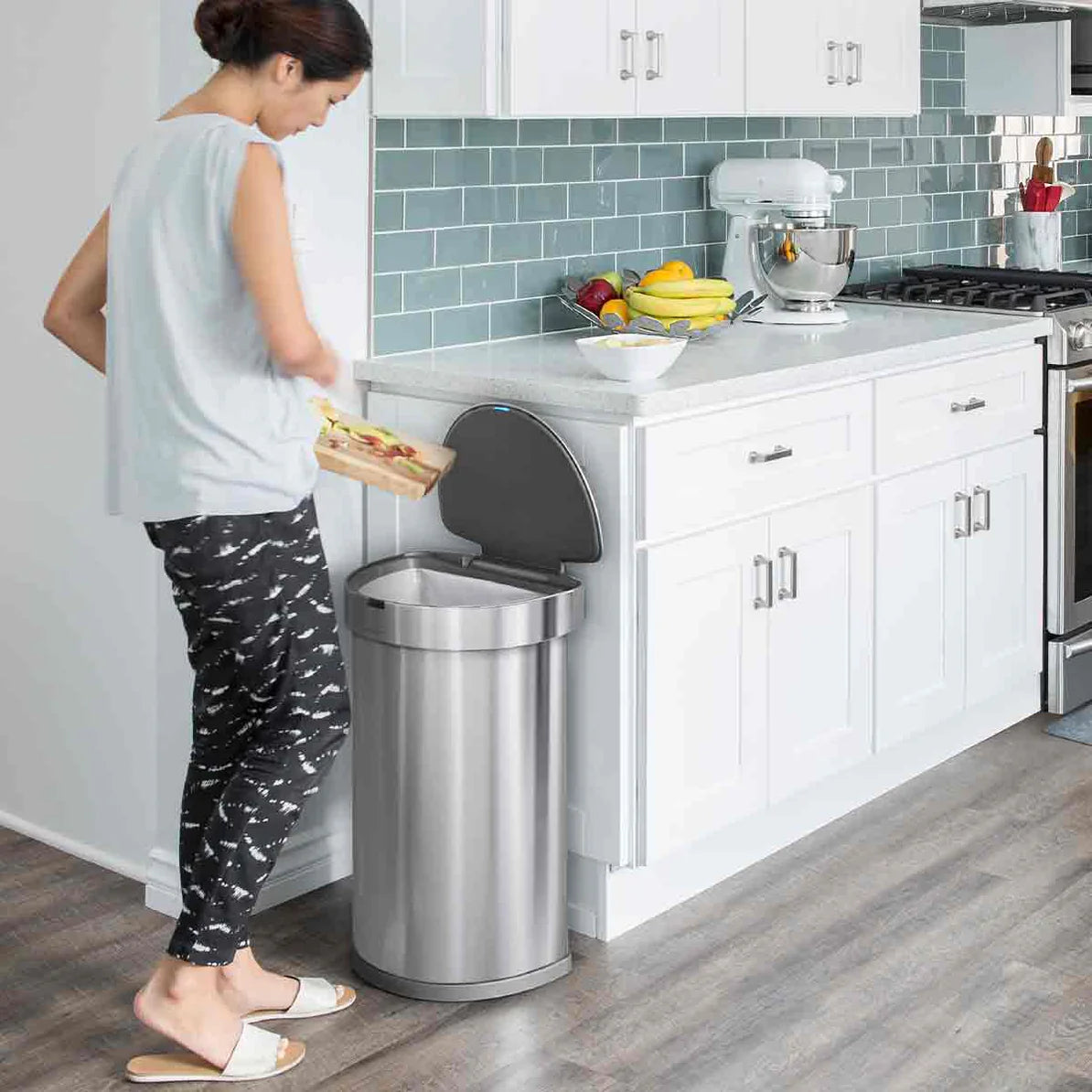 The image shows a kitchen setting with a woman discarding food into a Simplehuman stainless steel cylindrical trash can. The trash can is positioned against a kitchen counter with white cabinets and a blue-tiled backsplash. The can's lid is open, likely activated by motion, as the woman is about to dispose of a pizza box. The kitchen environment is modern and clean, with additional appliances and fruits visible on the counter, enhancing the overall contemporary aesthetic.