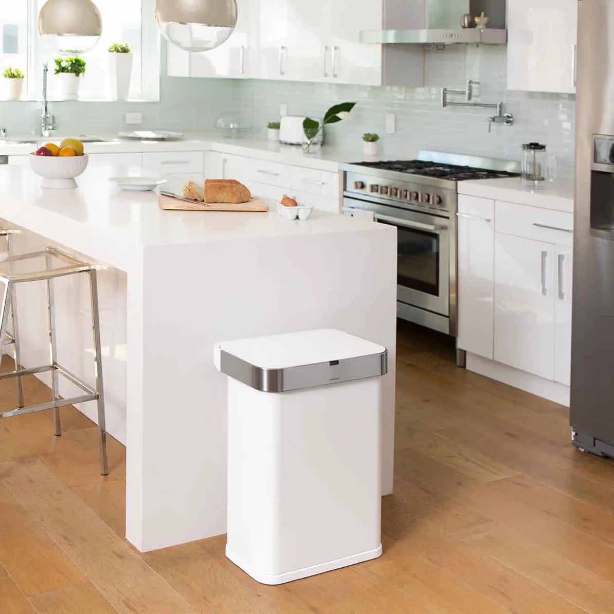 The image shows a modern, bright kitchen featuring a Simplehuman trash can with a white body and a stainless steel lid. The trash can is positioned near a kitchen island, blending seamlessly into the contemporary design of the space. The kitchen has a clean and minimalist aesthetic, with white cabinetry, stainless steel appliances, and a light-colored wooden floor.