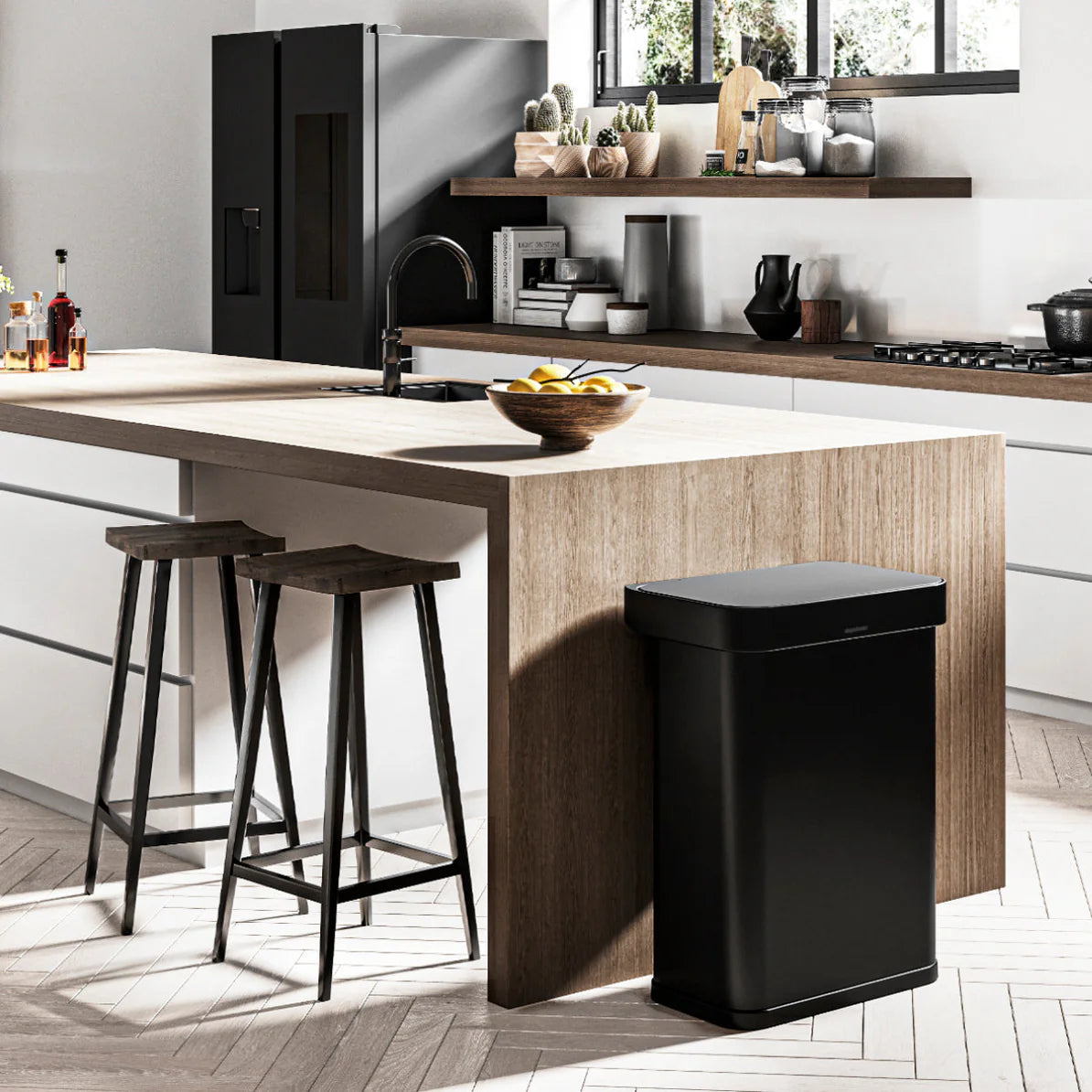 The image shows a modern kitchen interior featuring a Simplehuman trash can in matte black, placed at the end of a wooden kitchen island. The kitchen has a contemporary design with clean lines and a mix of natural and industrial elements. The island has a wood finish, and it is paired with two black bar stools, enhancing the minimalist and functional aesthetic of the space. On the island, there is a fruit bowl, adding a touch of natural color to the setting.