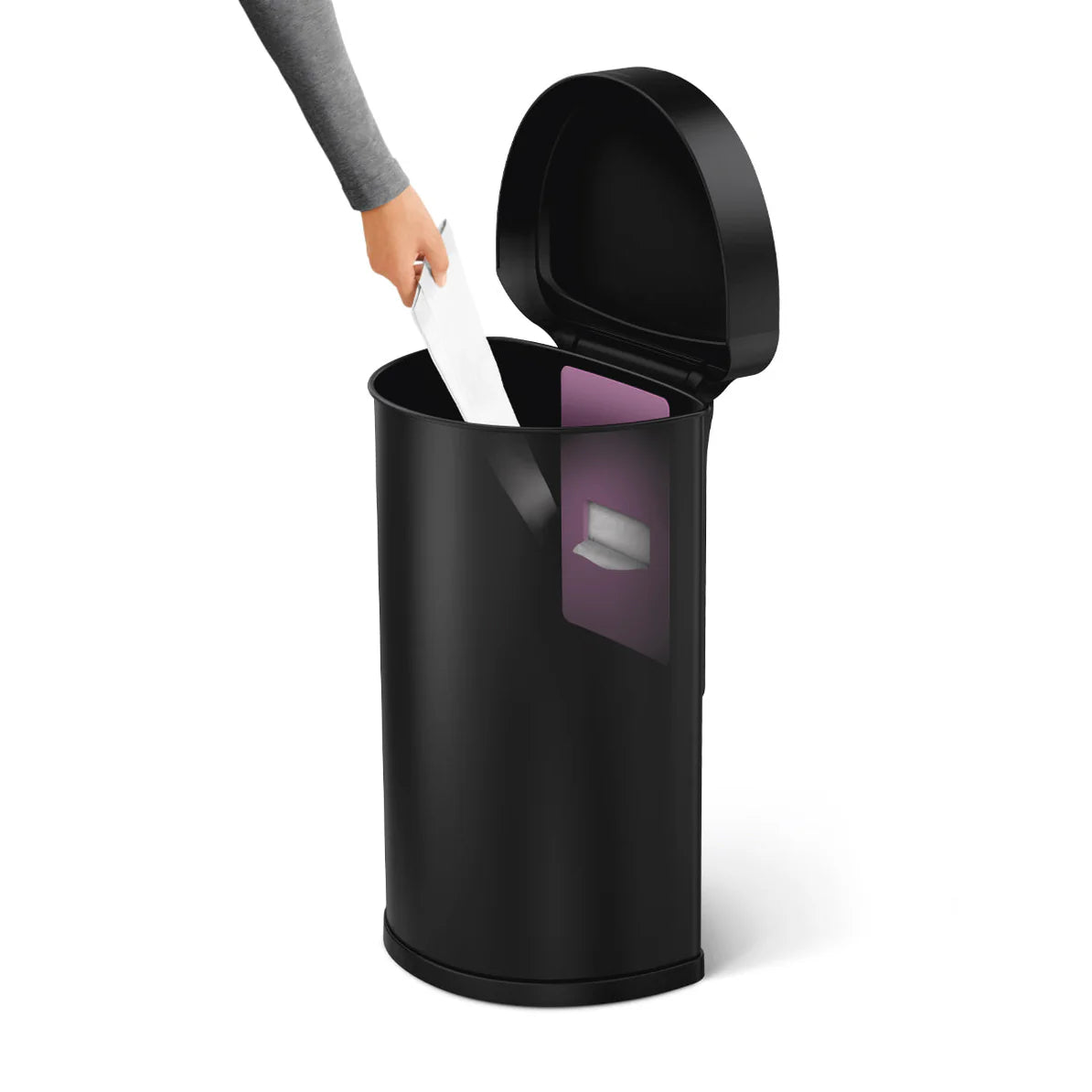 The image shows a Simplehuman trash can with a round design in black. The lid is open, revealing the interior of the trash can. A person’s hand is seen placing a white item (likely a trash bag or a piece of trash) into the can. Inside, there is a visible purple compartment, possibly for storing additional trash bags or for an odor-control feature. The can's sleek, modern design is consistent with Simplehuman's aesthetic, and the scene emphasizes the ease of use and functionality of the product.