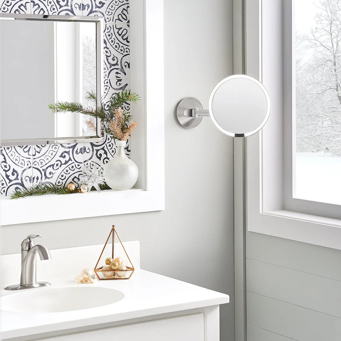 The image depicts a modern bathroom setting with a wall-mounted round mirror featuring a sleek silver frame and an adjustable arm. The mirror is positioned next to a window, allowing natural light to illuminate the space. The bathroom vanity area includes a white countertop with a silver faucet, complemented by decorative elements like a geometric gold holder with shells and a vase with greenery. The wall behind the mirror features a decorative patterned section, adding a stylish touch to the overall design