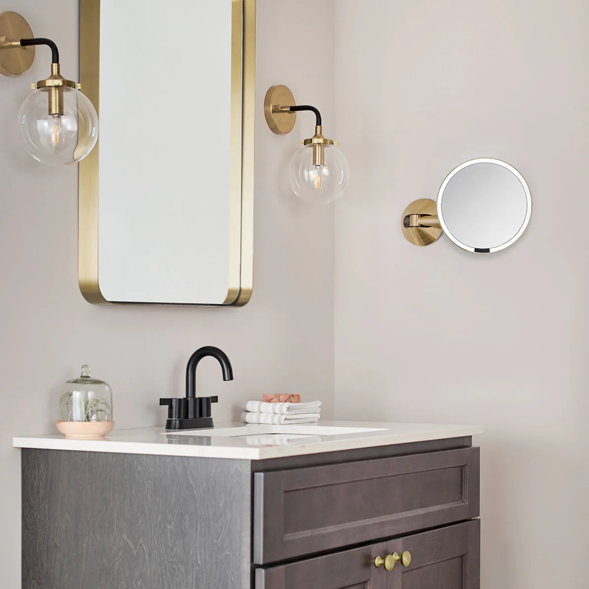 The image shows a stylish bathroom vanity setup featuring a wall-mounted round mirror with a gold frame and adjustable arm. The vanity area includes a dark wood cabinet with brass handles, a white countertop, and a sleek black faucet. Above the sink is a larger rectangular mirror with a matching gold frame. The space is illuminated by elegant wall sconces with glass globe shades and gold accents. The combination of warm gold tones, dark wood, and modern fixtures creates a luxurious and contemporary bathroom