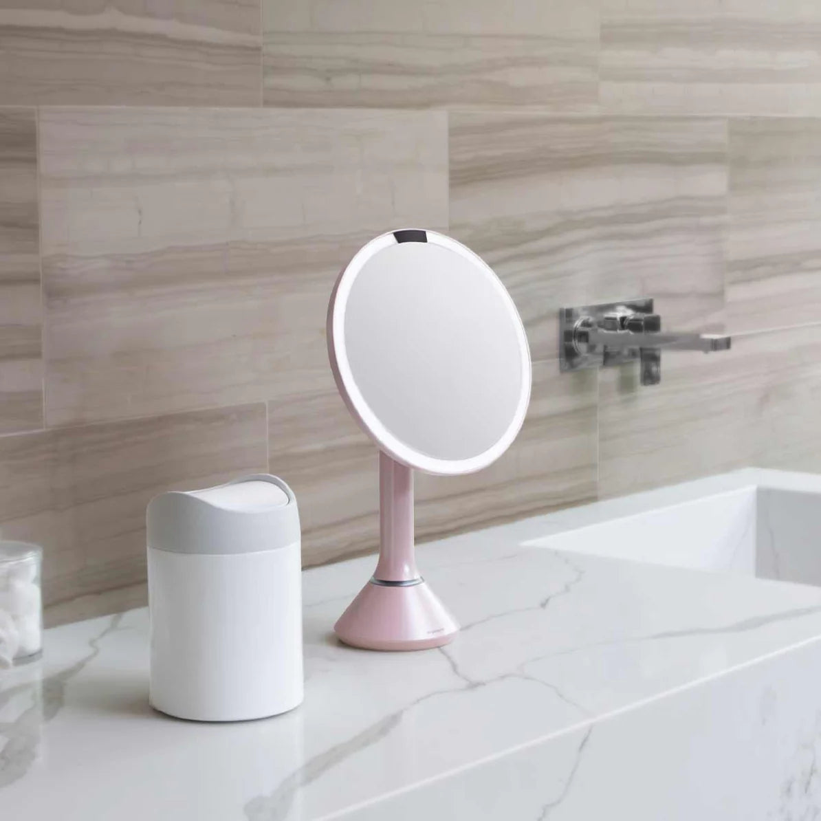 This image shows a Simplehuman makeup mirror with a soft pink finish, placed on a marble countertop in a bathroom. The mirror's LED light ring is visible, providing clear and bright illumination for precise makeup application or grooming. The pink color adds a delicate, feminine touch to the mirror, making it a stylish accessory that can enhance the aesthetic of any bathroom or vanity space.  Next to the mirror is a sleek, white container with a modern design, possibly a storage bin or another bathroom acce