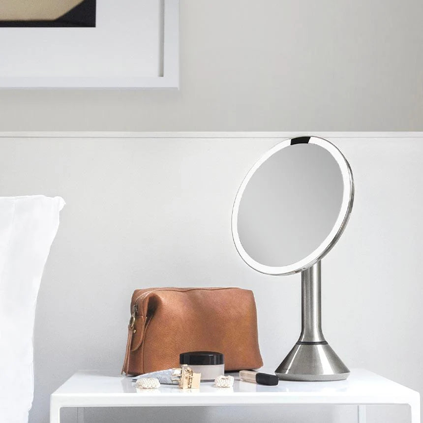 This image displays a Simplehuman makeup mirror with a stainless steel finish, placed on a white vanity or tabletop. The mirror's LED light ring is prominent, suggesting that it provides bright, even illumination, ideal for makeup application or grooming.  Next to the mirror is a stylish brown leather makeup bag, along with various beauty products, including a jar of cream, lipstick, and some accessories. These items are neatly arranged, creating a tidy and elegant display that highlights the mirror as part