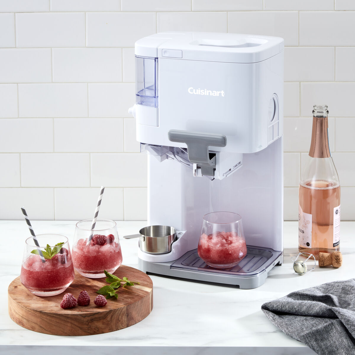 Cuisinart soft serve machine used to prepare raspberry slush drinks, with glasses of slush garnished with mint and raspberries beside a bottle of rosé.