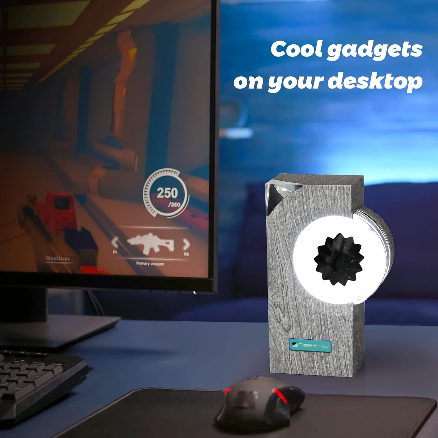 The image shows a ferrofluid music companion placed on a desktop beside a gaming setup. The text "Cool gadgets on your desktop" emphasizes its role as a stylish and interactive accessory for tech enthusiasts. The device's dynamic ferrofluid display adds a unique visual element to the workspace, making it a perfect companion for those who appreciate innovative gadgets while gaming or working.