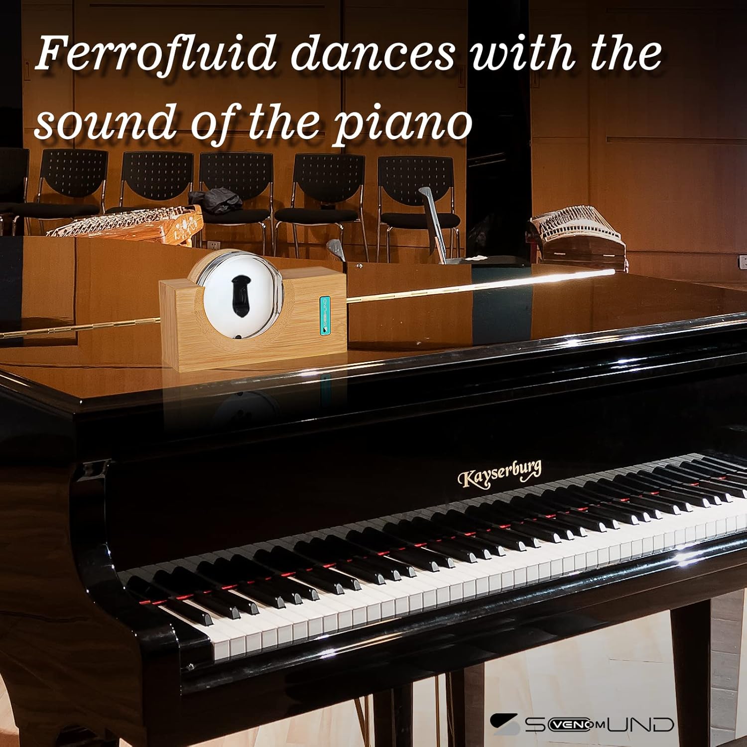 A ferrofluid magnetic liquid audio display is placed on a sleek black Kayserburg piano. The image caption highlights the interaction between the ferrofluid and the piano's music, noting how the ferrofluid "dances" with the sound. The setting appears to be in a concert hall or performance space, with the ferrofluid reacting to the piano's notes in a visually captivating way. This image emphasizes the artistic fusion of sound and science.