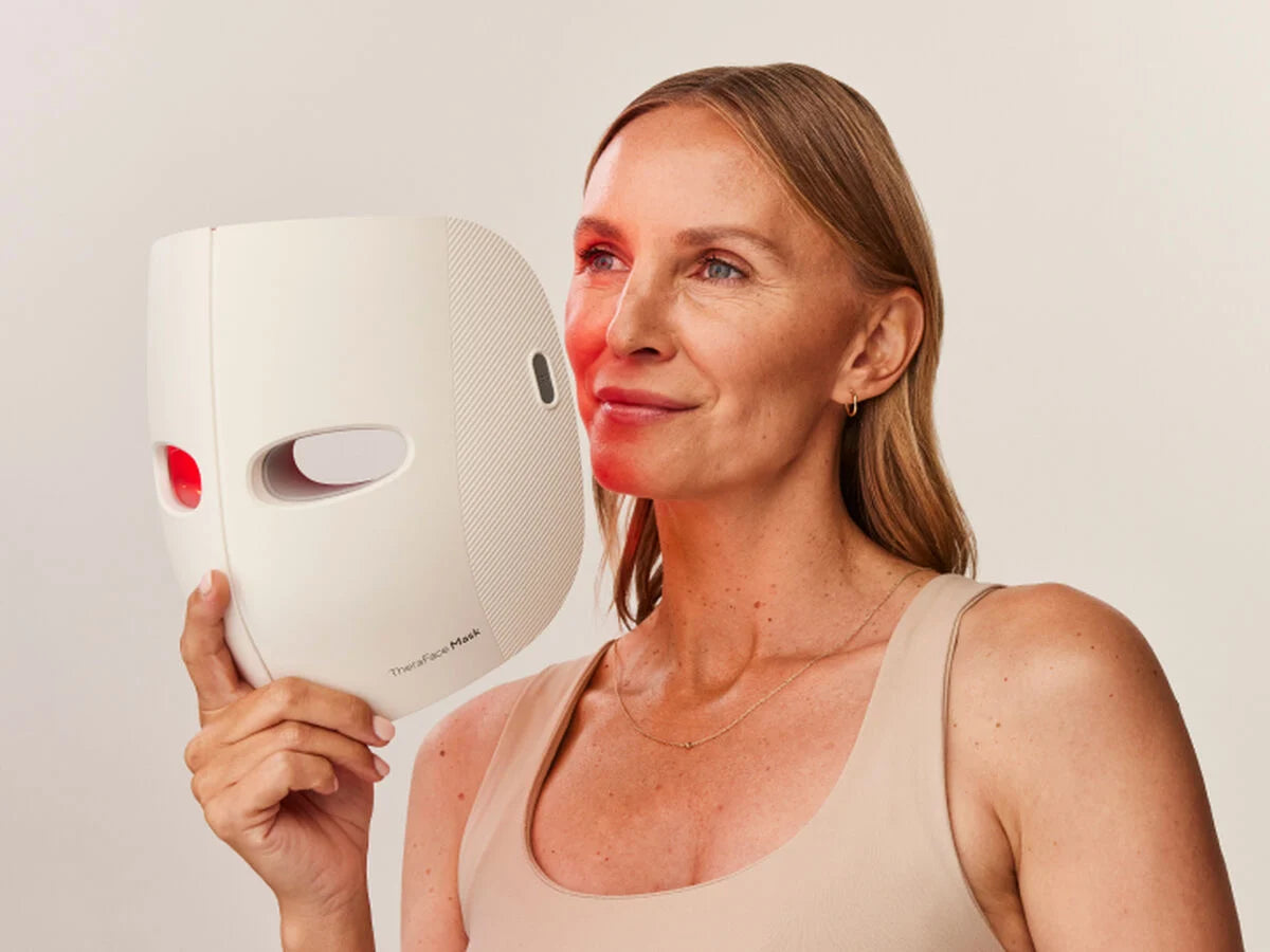 A woman holding a white LED therapy face mask close to her face, with red light emitting from the mask onto her skin. She is wearing a beige tank top and has a relaxed, confident expression. The mask, labeled "Thea Face Mask," is designed for skincare treatments to improve skin health and radiance. The background is a neutral, minimalistic setting.