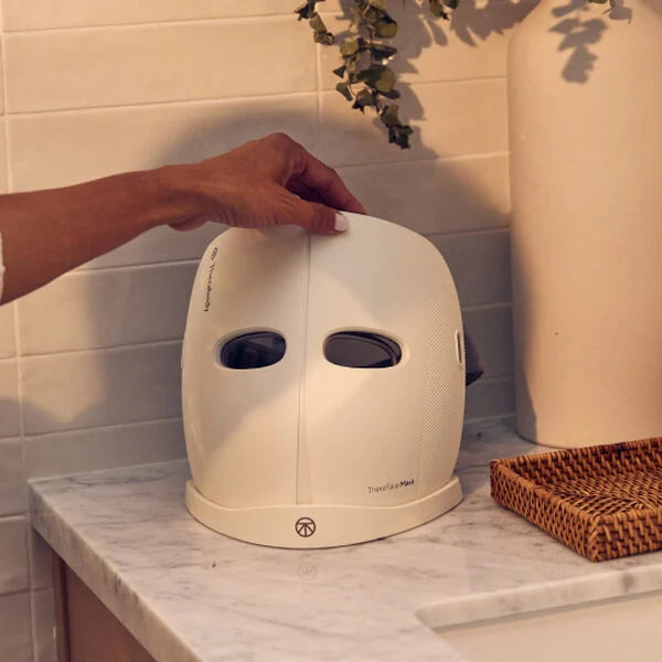 A person placing a white LED therapy face mask on a marble countertop. The mask features a sleek design with eye cutouts and is labeled "Thea Face Mask" on the front. The setting includes a woven basket and a decorative vase in the background, creating a spa-like ambiance. The mask is designed for skincare treatments, offering benefits such as improving skin tone and reducing blemishes.