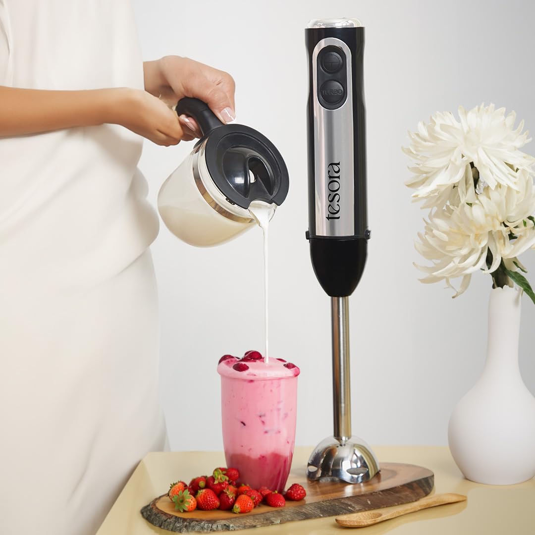 This image showcases the **Tesora hand blender** being used to prepare a delicious smoothie or shake. The blender is placed beside a serving of the freshly made pink drink, complemented with strawberries and cream. It highlights the hand blender’s versatility for preparing thick, creamy beverages quickly and efficiently. The design of the blender, with its sleek look and stainless steel blending arm, adds to its appeal as a reliable kitchen companion for blending smoothies, shakes, and more.