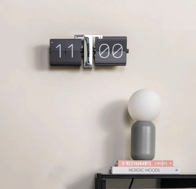 Stylish flip clock displaying "11:00" mounted on a beige wall above a sleek black shelf. The shelf holds a modern gray lamp with a round white bulb and stacked books titled "Restaurants" and "Nordic Moods," creating a sophisticated and minimalist decor scene.