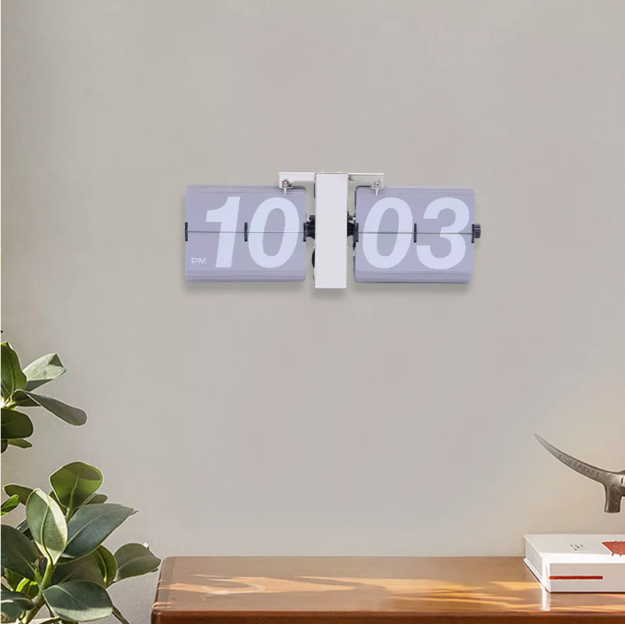 Minimalist flip clock displaying "10:03 PM" mounted on a neutral wall, set above a wooden surface with a few plants and decor items, adding a modern and clean aesthetic to the space.