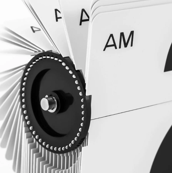 Close-up of a mechanical flip clock mechanism, highlighting the rotating black gear and white panels displaying "AM," emphasizing the intricate details and precision of the timekeeping design.