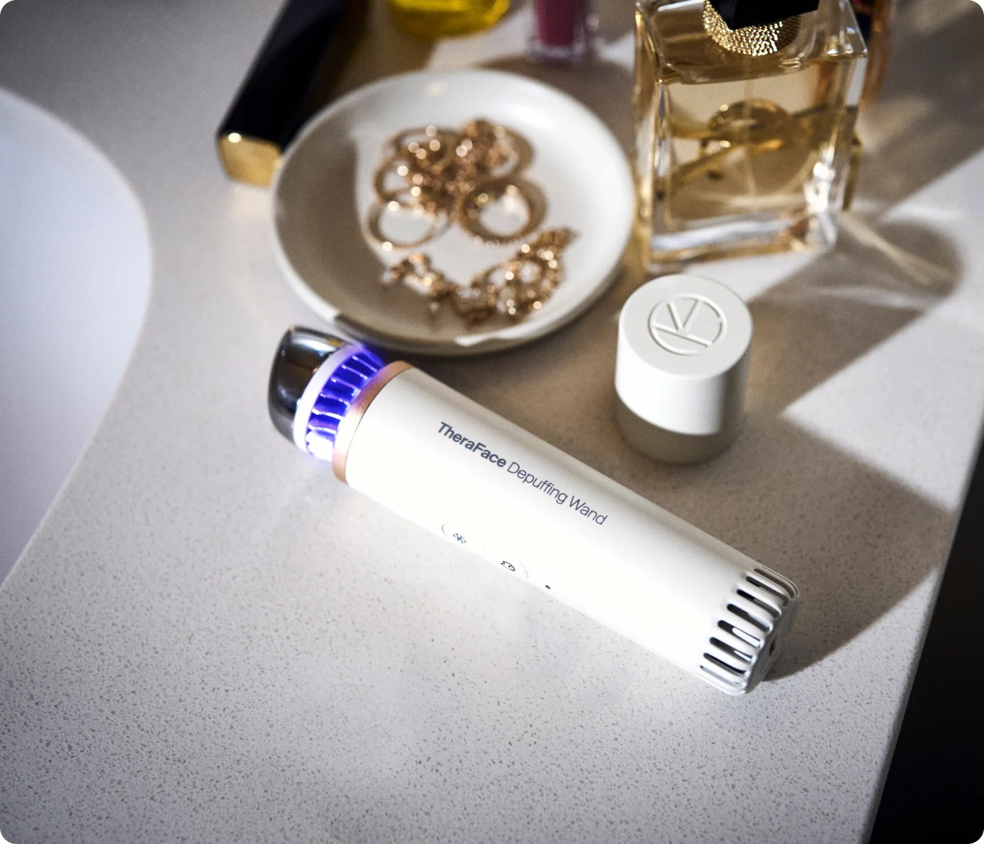 White TheraFace Depuffing Wand on a vanity with the blue cold-treatment LED lit, beside jewellery and perfume