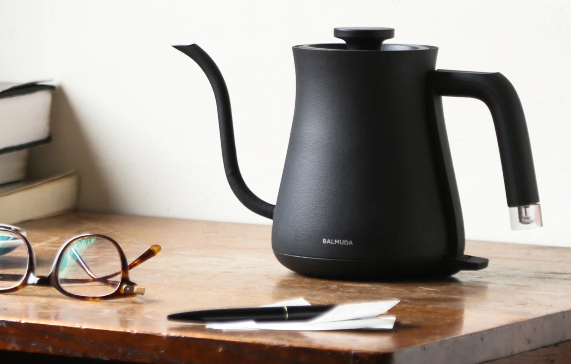 The image shows a Balmuda electric kettle in a matte black finish placed on a wooden desk or tabletop. The kettle features a minimalist design with a curved spout and an ergonomic handle. Nearby on the table are a pair of tortoiseshell glasses, a pen, and a few stacked books, creating a cozy and studious atmosphere.