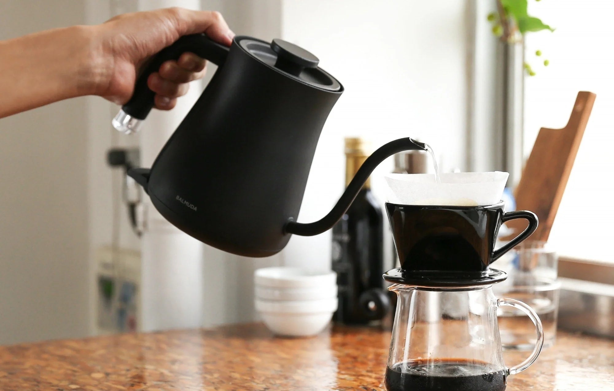 The image shows a person pouring hot water from a Balmuda electric kettle in a matte black finish into a pour-over coffee dripper set on top of a glass carafe. The kettle has a sleek design with a curved spout for precise pouring and an ergonomic handle