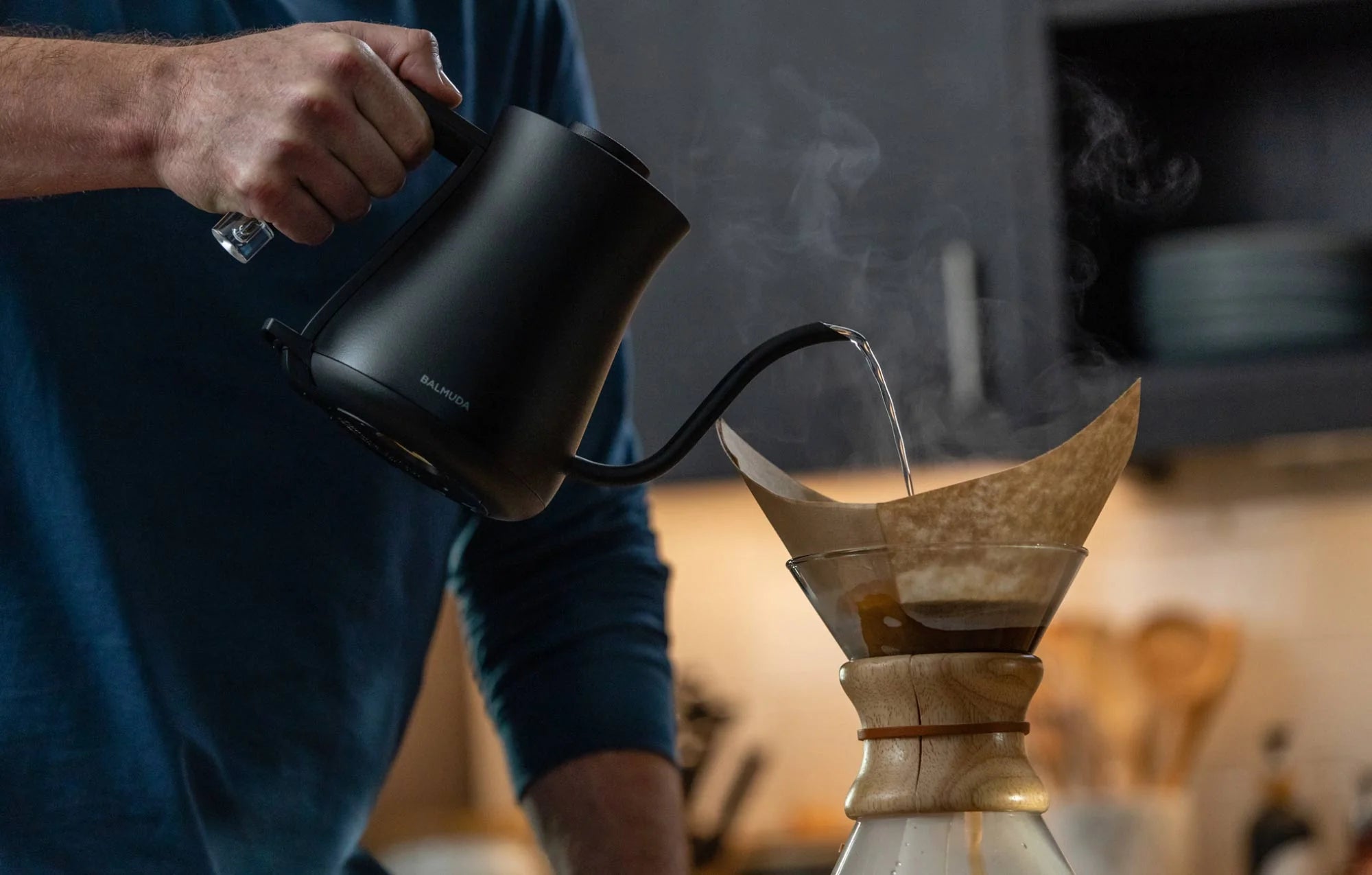 The image shows a person pouring hot water from a Balmuda electric kettle in a matte black finish into a Chemex coffee maker. The kettle features a long, curved spout for precise pouring, and steam is visible rising from the kettle and the coffee maker.