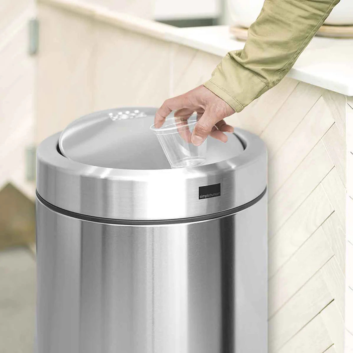 The image features a sleek, stainless steel trash can situated in a modern kitchen setting. The trash can has a black lid and base, complemented by a foot pedal for hands-free operation. It's positioned near stainless steel appliances and wooden cabinetry, blending seamlessly with the contemporary decor. The trash can's polished finish reflects the surrounding elements, emphasizing its stylish and functional design within the kitchen space.