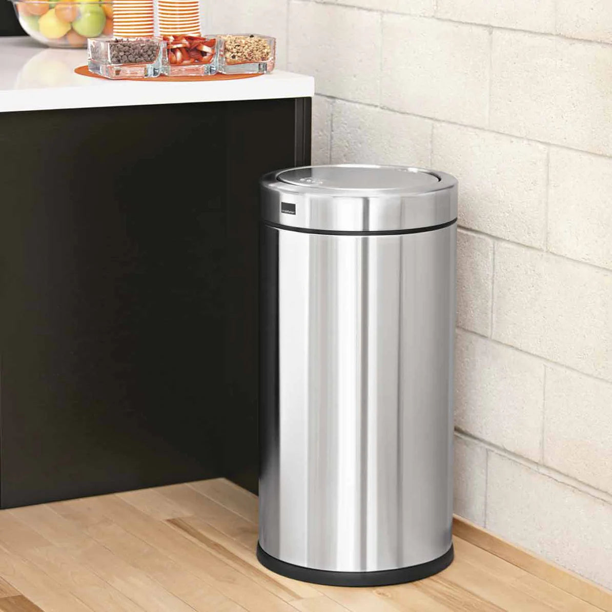 The image features a sleek, cylindrical stainless steel trash can positioned in a modern kitchen setting. The trash can has a flat, circular lid and a black base, providing a clean and contemporary look. Its polished stainless steel finish complements the surrounding decor, making it both a functional and stylish addition to the space. The trash can's design is ideal for fitting neatly into corners or against walls, offering efficient waste management without compromising on aesthetics.