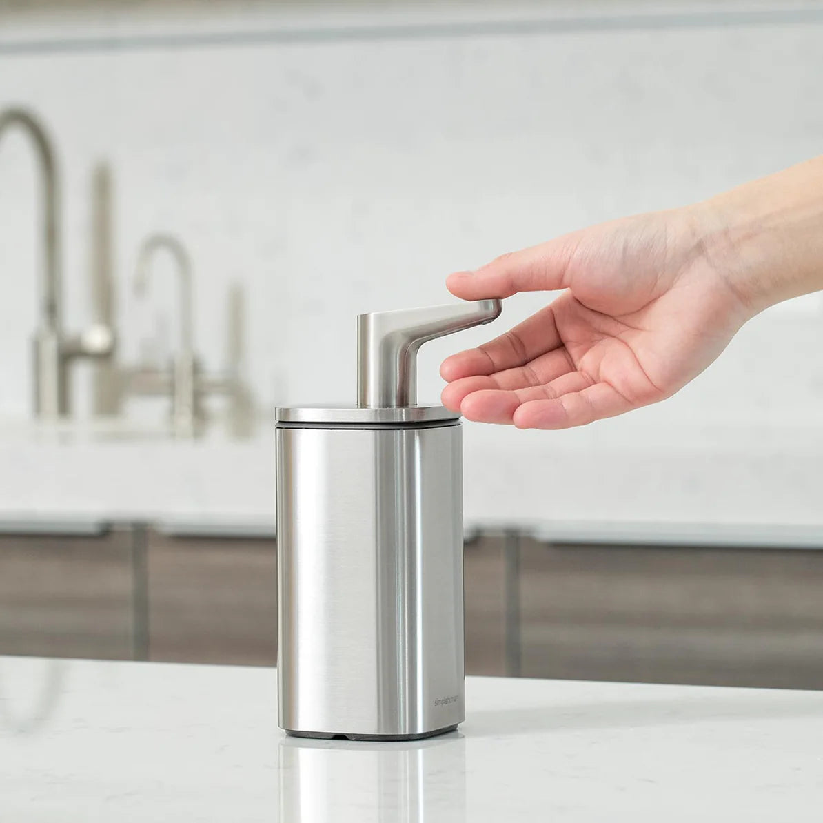 The image shows a sleek, silver automatic soap dispenser in a modern kitchen setting. A hand is positioned under the dispenser's nozzle, likely to trigger the sensor for soap dispensing. The dispenser has a metallic finish with a clean, minimalist design, characteristic of Simplehuman products. The kitchen environment in the background emphasizes the dispenser's suitability for contemporary home use, blending both functionality and aesthetic appeal.