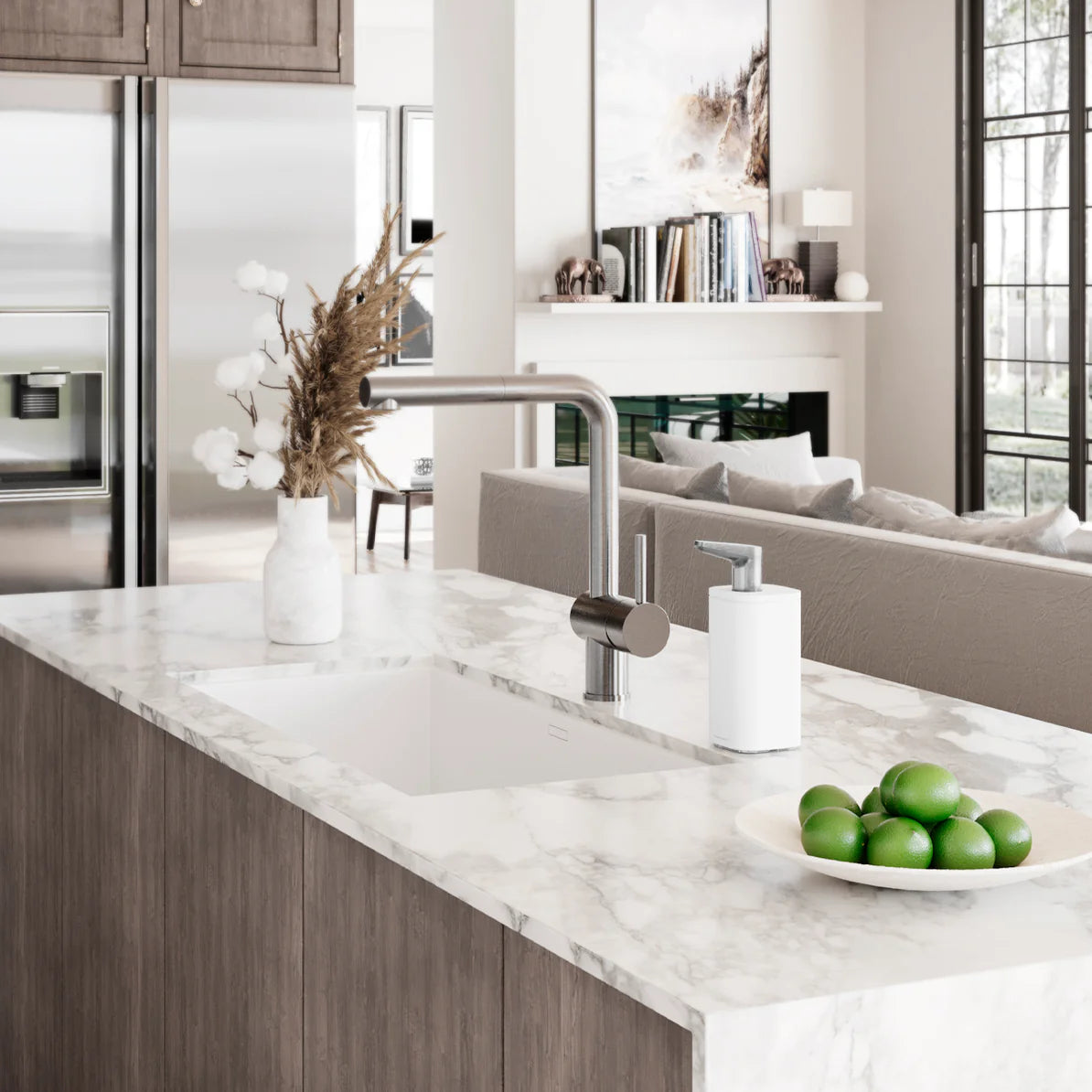 The image shows a modern kitchen setting with a marble countertop and a sleek, minimalist design. On the countertop, there is a white automatic soap dispenser placed next to the sink, blending seamlessly with the elegant decor. The kitchen features stainless steel appliances, a stylish faucet, and a bowl of limes adding a pop of color. In the background, the open-concept living space is visible, with a comfortable sofa, a fireplace, and large windows that allow natural light to fill the room. The overall ae