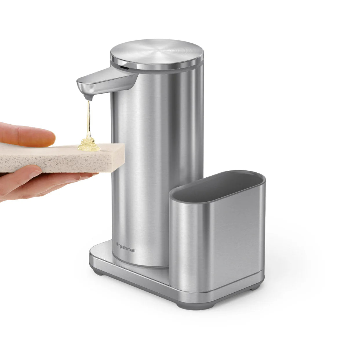 A stainless steel automatic soap dispenser dispensing soap onto a sponge being held by a hand. The dispenser is cylindrical and stands on a rectangular base with an adjacent sponge holder. The soap is being dispensed from a nozzle near the top of the dispenser.