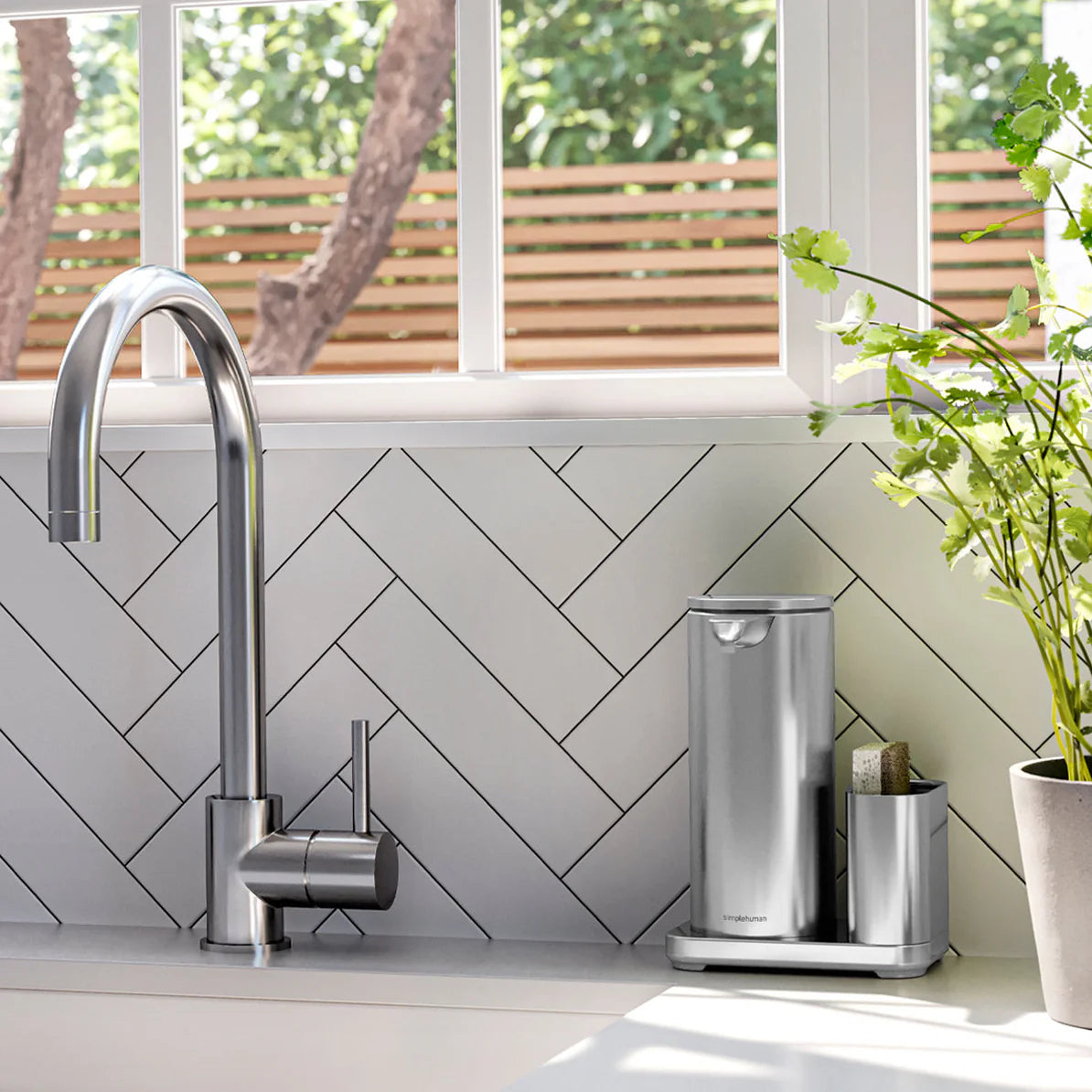 A modern kitchen sink area featuring a stainless steel automatic soap dispenser and sponge holder set on a countertop next to a potted plant. The dispenser and holder are positioned against a white herringbone tile backsplash. A sleek, curved faucet is installed to the left of the soap dispenser, with a window and wooden fence visible in the background.