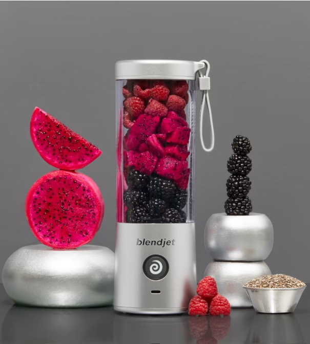 BlendJet portable blender filled with vibrant fruits like dragon fruit, raspberries, and blackberries, surrounded by artistic arrangements of sliced dragon fruit, stacked blackberries, fresh raspberries, and a bowl of chia seeds, set against a sleek gray background.