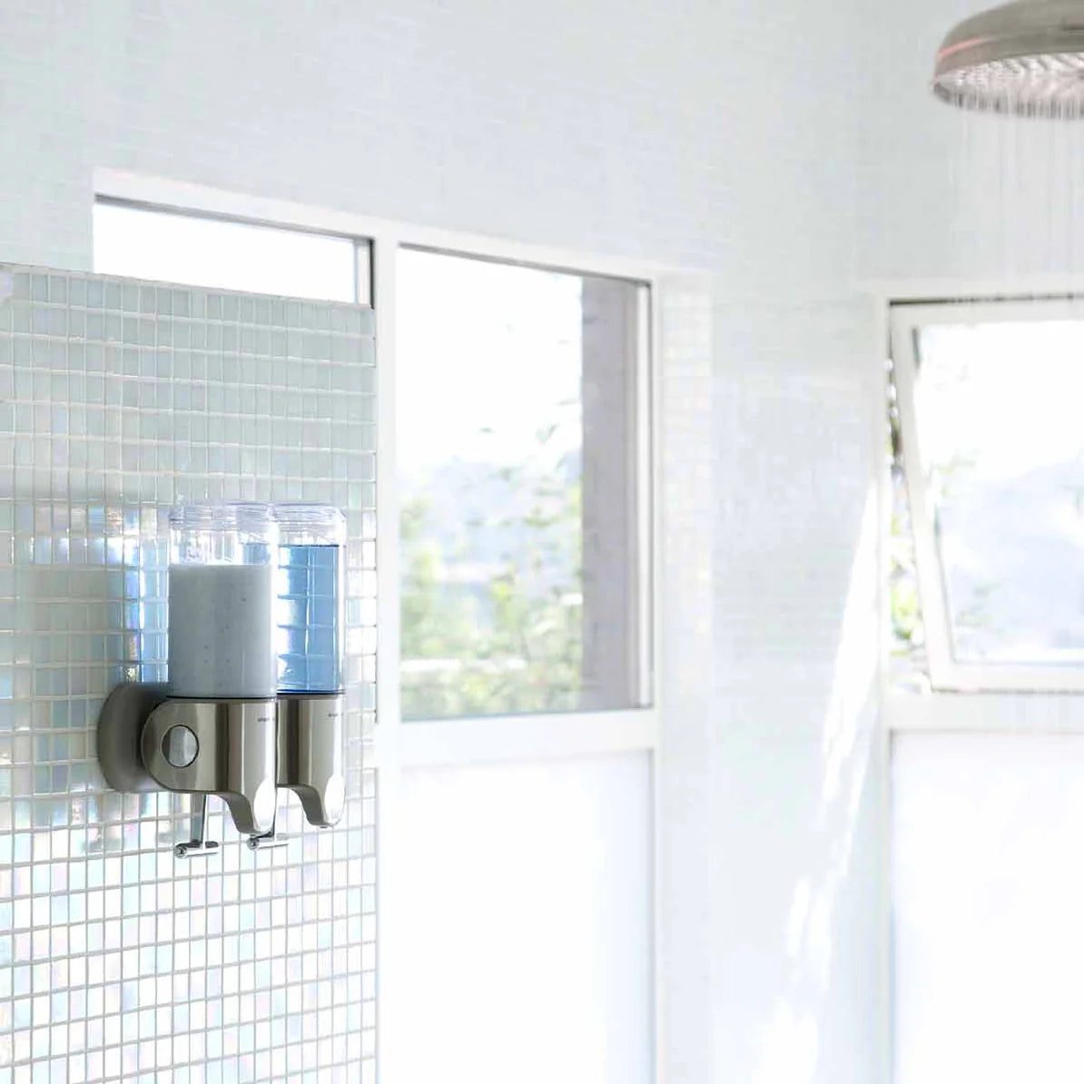 The image shows a modern bathroom setting with a wall-mounted dual dispenser system. The dispensers are attached to a mosaic tile wall and contain different liquids, one light gray and the other light blue, likely used for shampoo and conditioner or other bathroom essentials. The dispensers have a stainless steel base with clear plastic containers, providing a sleek and contemporary look that complements the bright and airy atmosphere of the bathroom. Large windows allow natural light to flood the space, en