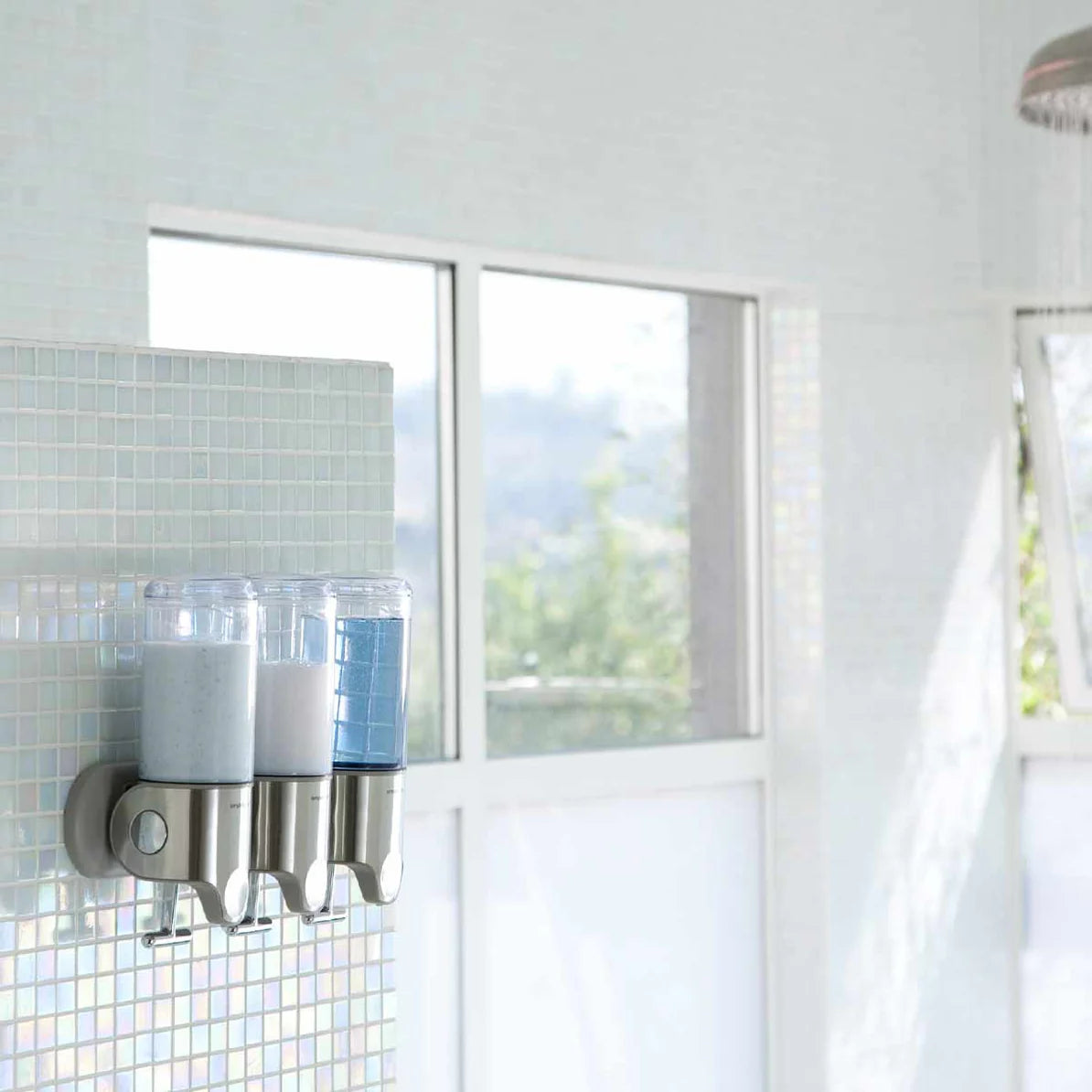 The image shows a bathroom setting with a wall-mounted triple dispenser system. The dispensers are filled with different liquids—likely shampoo, conditioner, and body wash, indicated by the colors in the containers (one light blue, one white, and one light gray). The dispensers are made of stainless steel or a similar material and have a sleek, modern design that complements the light-colored mosaic tile wall they are mounted on. The bathroom has large windows that allow natural light to flood the space, cr